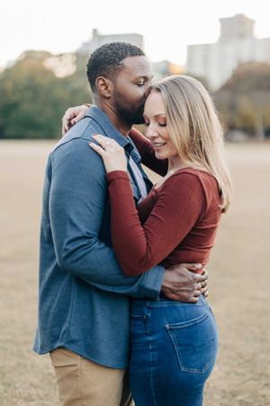 Interracial couple sharing a tender embrace outdoors with a city skyline visible in the background.