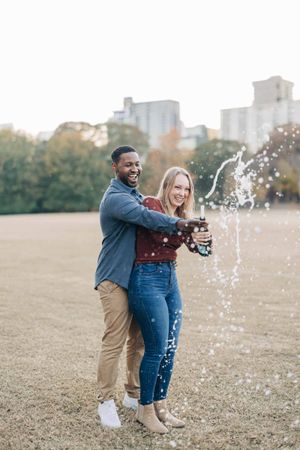 Happy interracial couple celebrating outdoors, popping champagne bottle in a sunny park with city buildings in background.