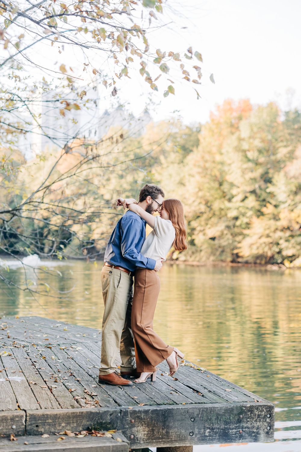 Couple embracing on a wooden dock by a serene lake surrounded by colorful autumn foliage.