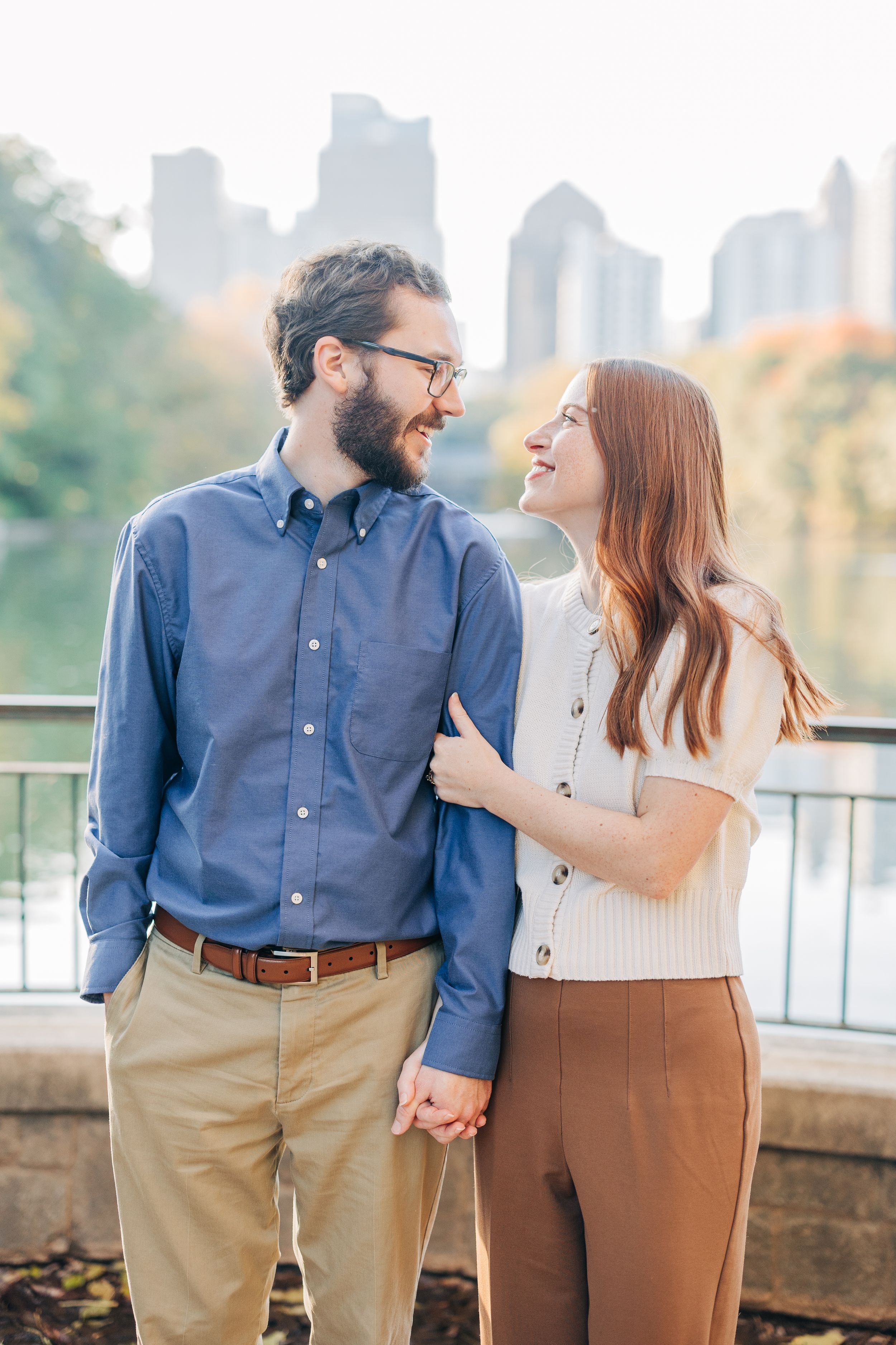 Happy couple holding hands and smiling at each other in Central Park with NYC skyline in the background.