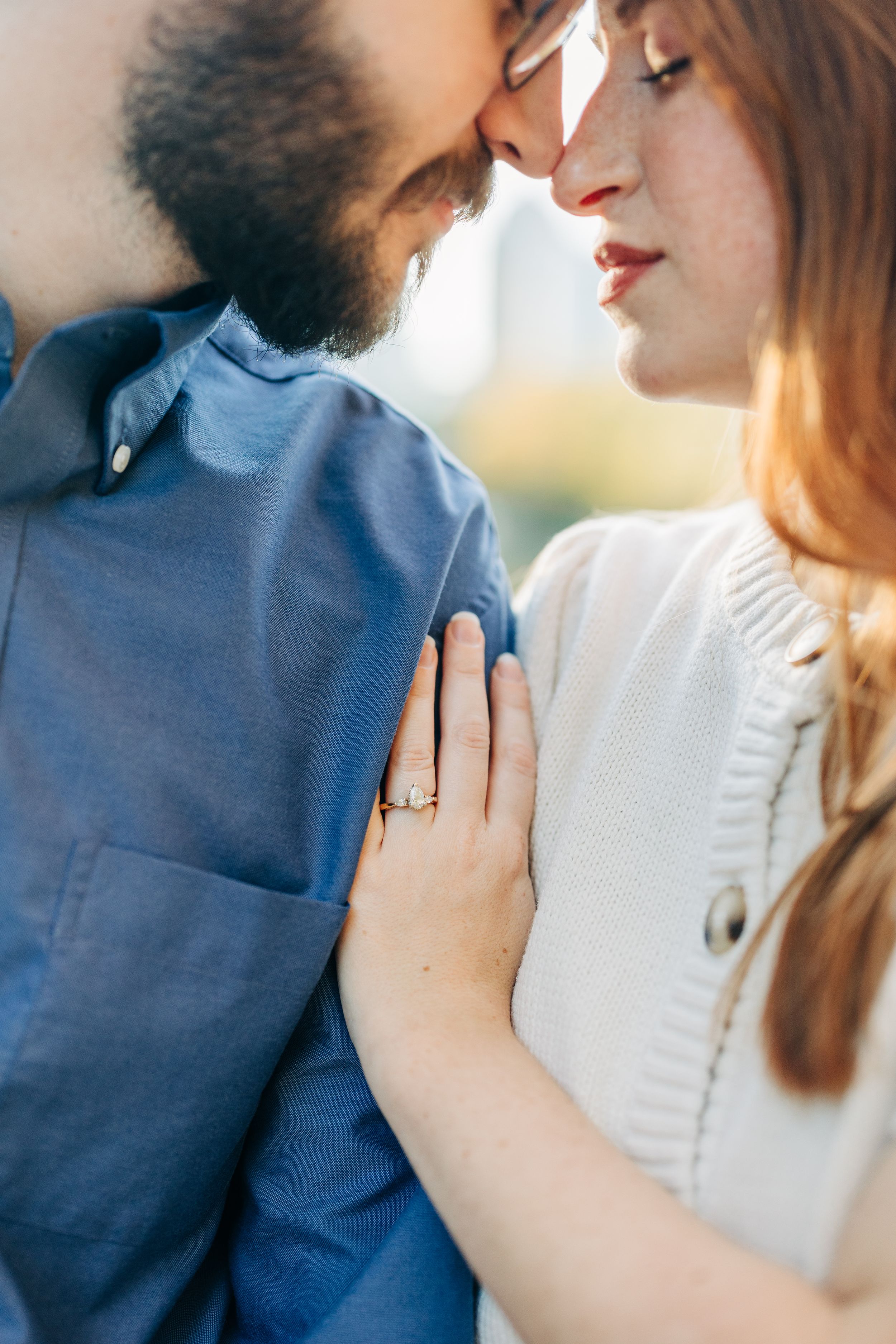 Couple sharing a romantic kiss, woman wearing an engagement ring resting her hand on man's blue shirt.