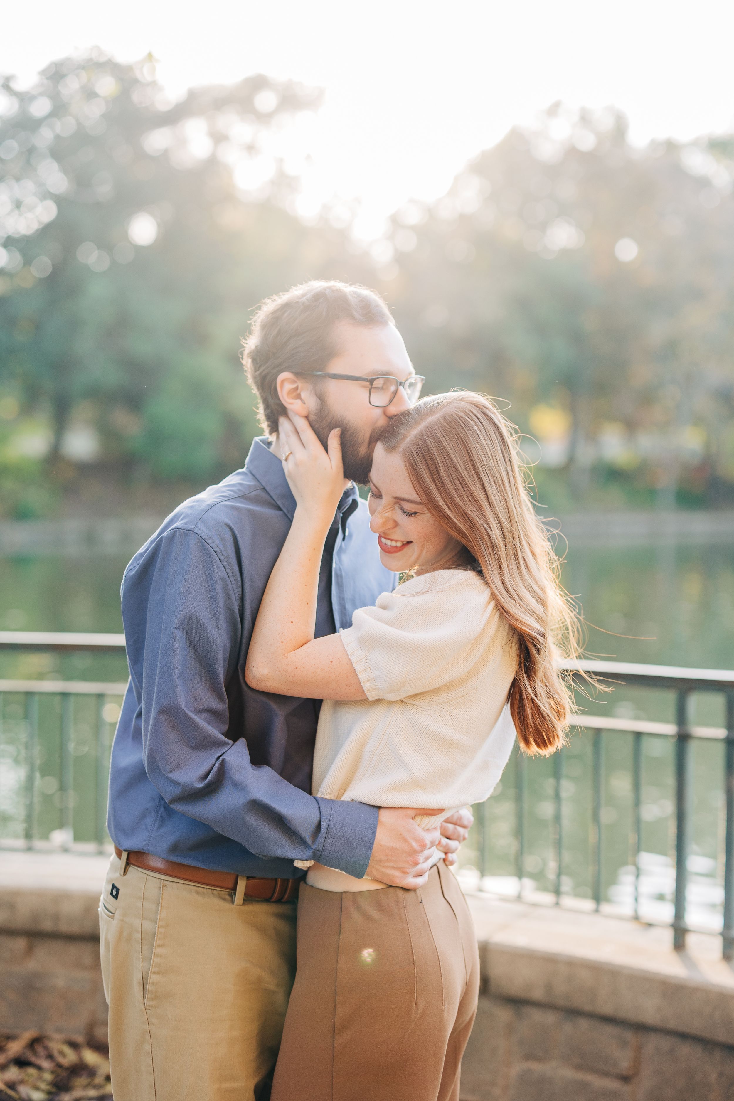 A couple embraces and laughs together near a sunlit pond, the man wearing glasses and a blue shirt, the woman in a cream top.