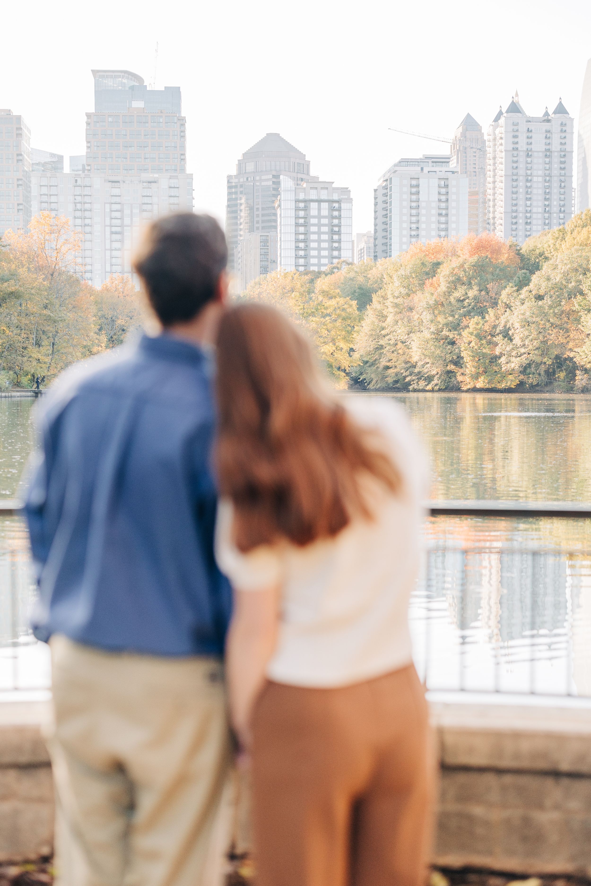 Couple standing together overlooking a tranquil lake with autumn trees and city skyline in the background.