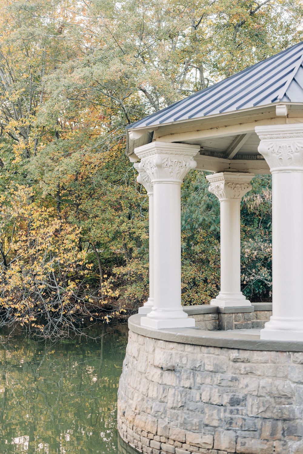 White classical gazebo with ornate columns sits on stone base beside tranquil pond surrounded by autumn foliage.