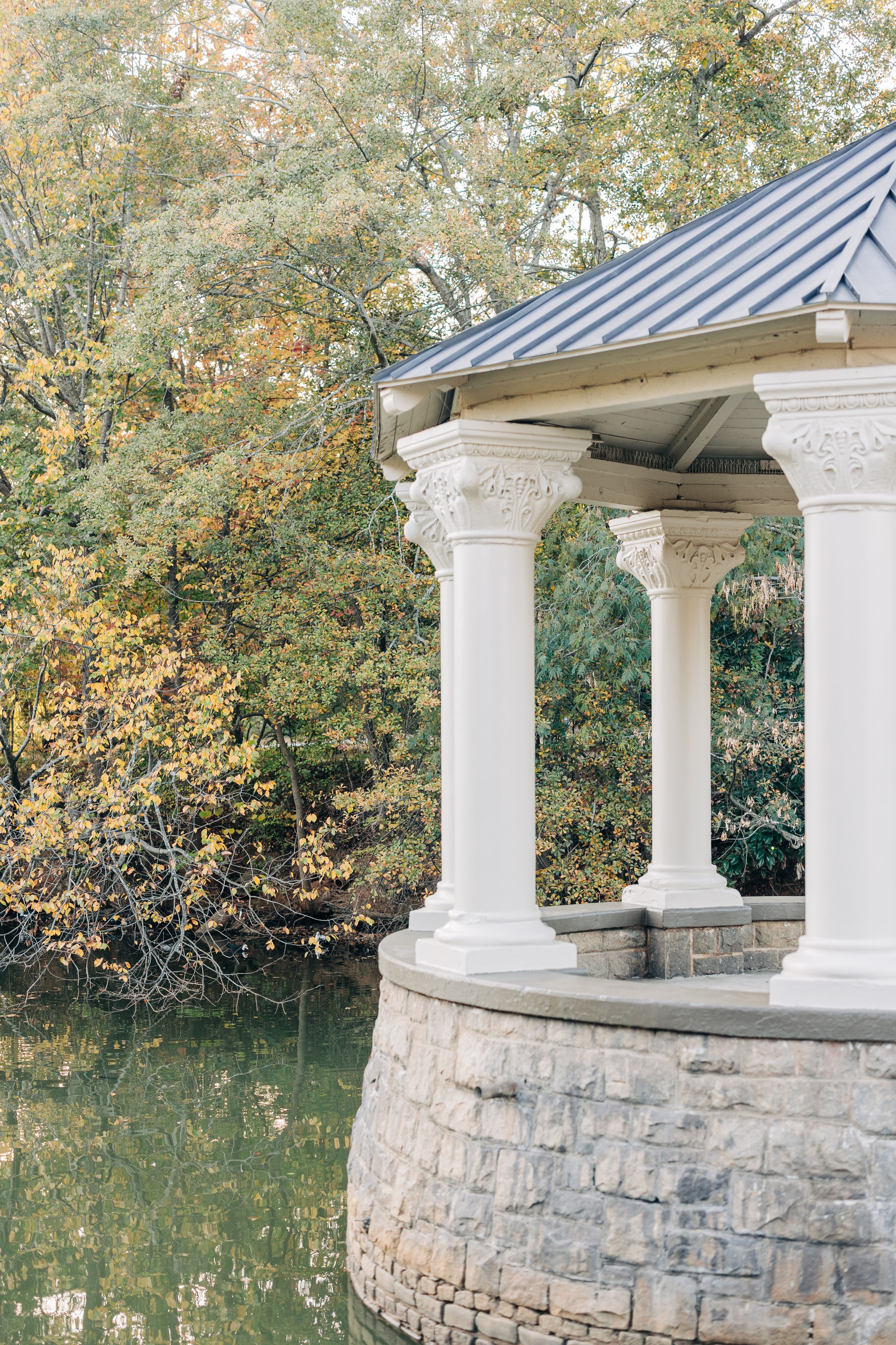 White classical gazebo with ornate columns sits on stone base beside tranquil pond surrounded by autumn foliage.