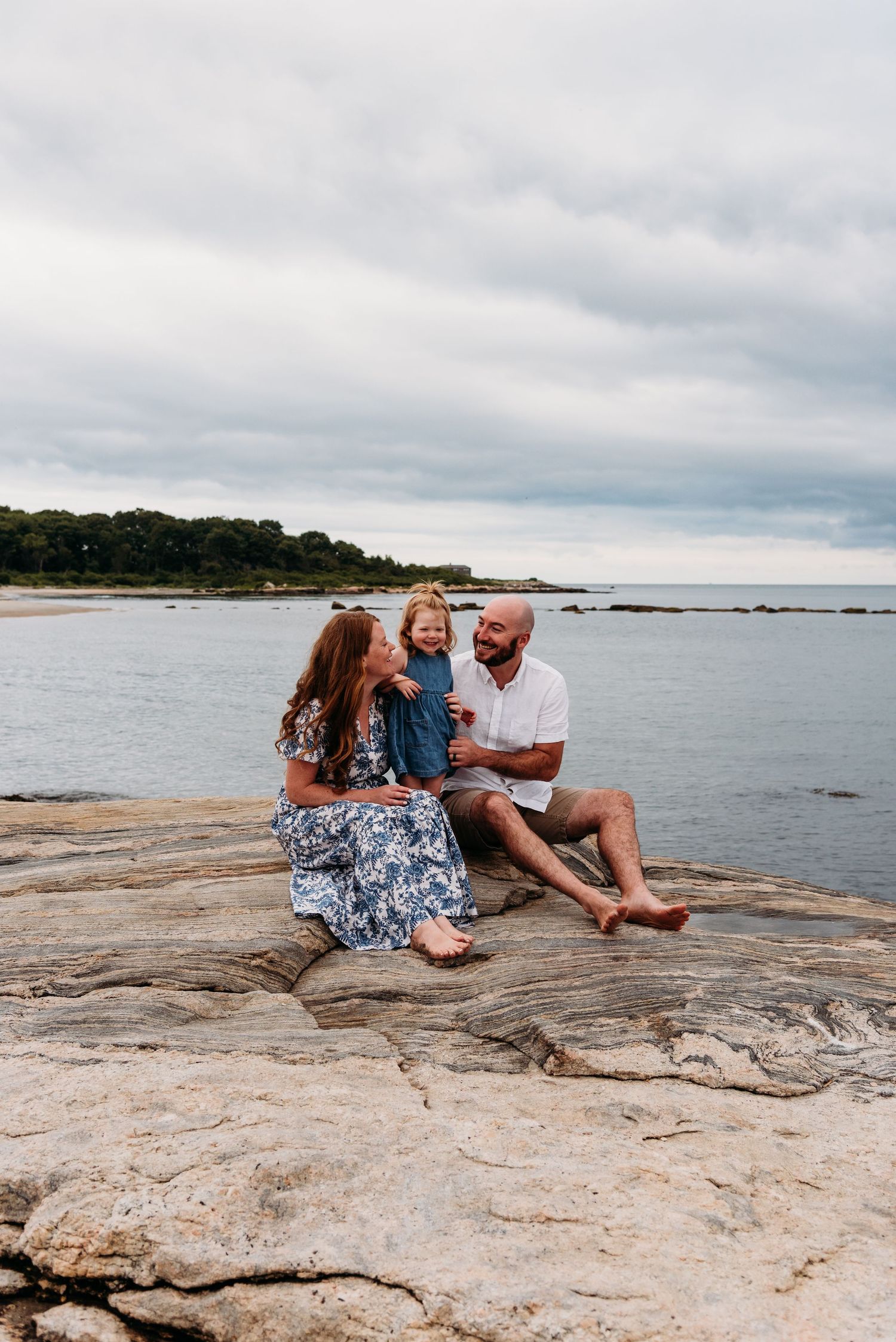 A family enjoys a peaceful moment together sitting on rocky coastal outcrop by the ocean under cloudy skies.
