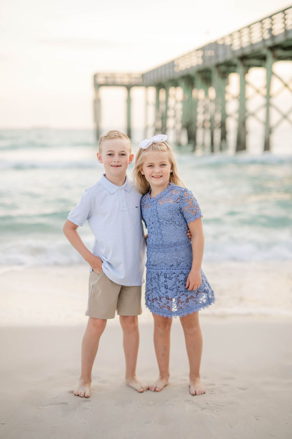 Two children standing barefoot on a beach by a pier at sunset wearing light blue and khaki casual attire.