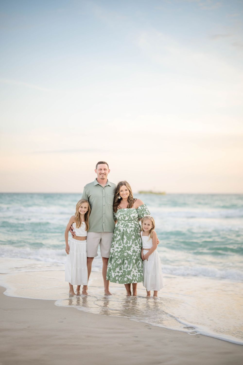 Family standing together at sunset on a beach with turquoise ocean waves and soft sandy shore in the background.