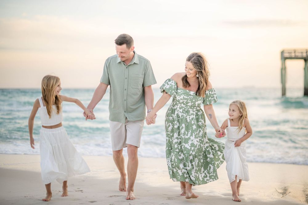 A family walks together hand in hand along a sandy beach at sunset with an ocean pier in the background.