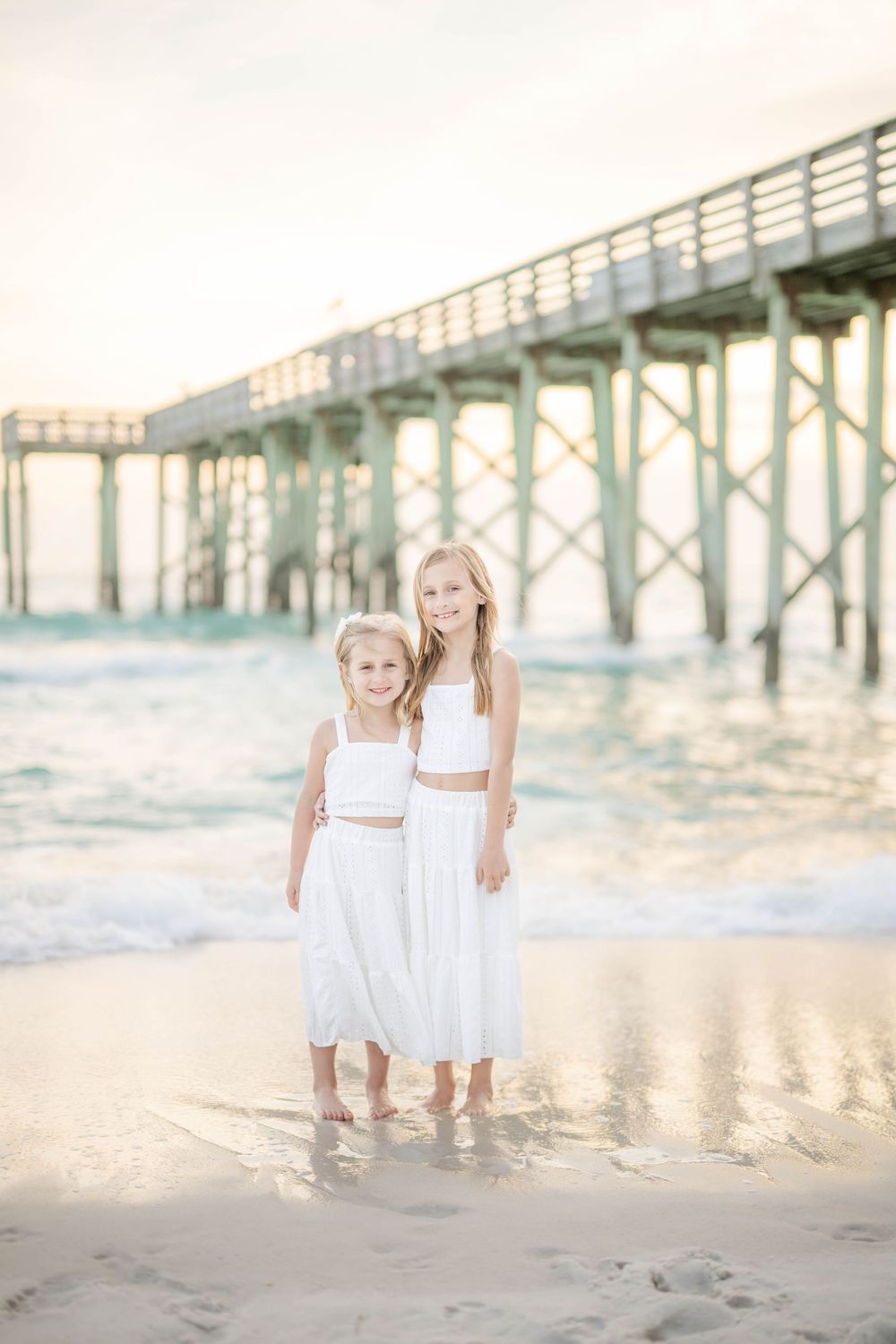 Ethereal beach scene with white dresses against a wooden pier at sunset, soft pastel colors reflecting on the sand.