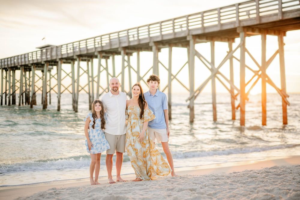 A group stands together on a beach at sunset with a wooden pier in the background.