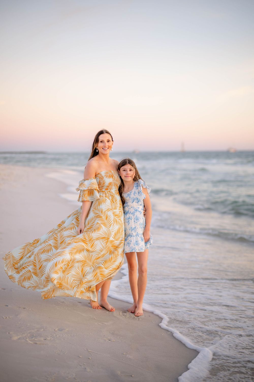 Two people in flowing dresses walk along a peaceful beach at sunset as waves gently roll onto the shore.