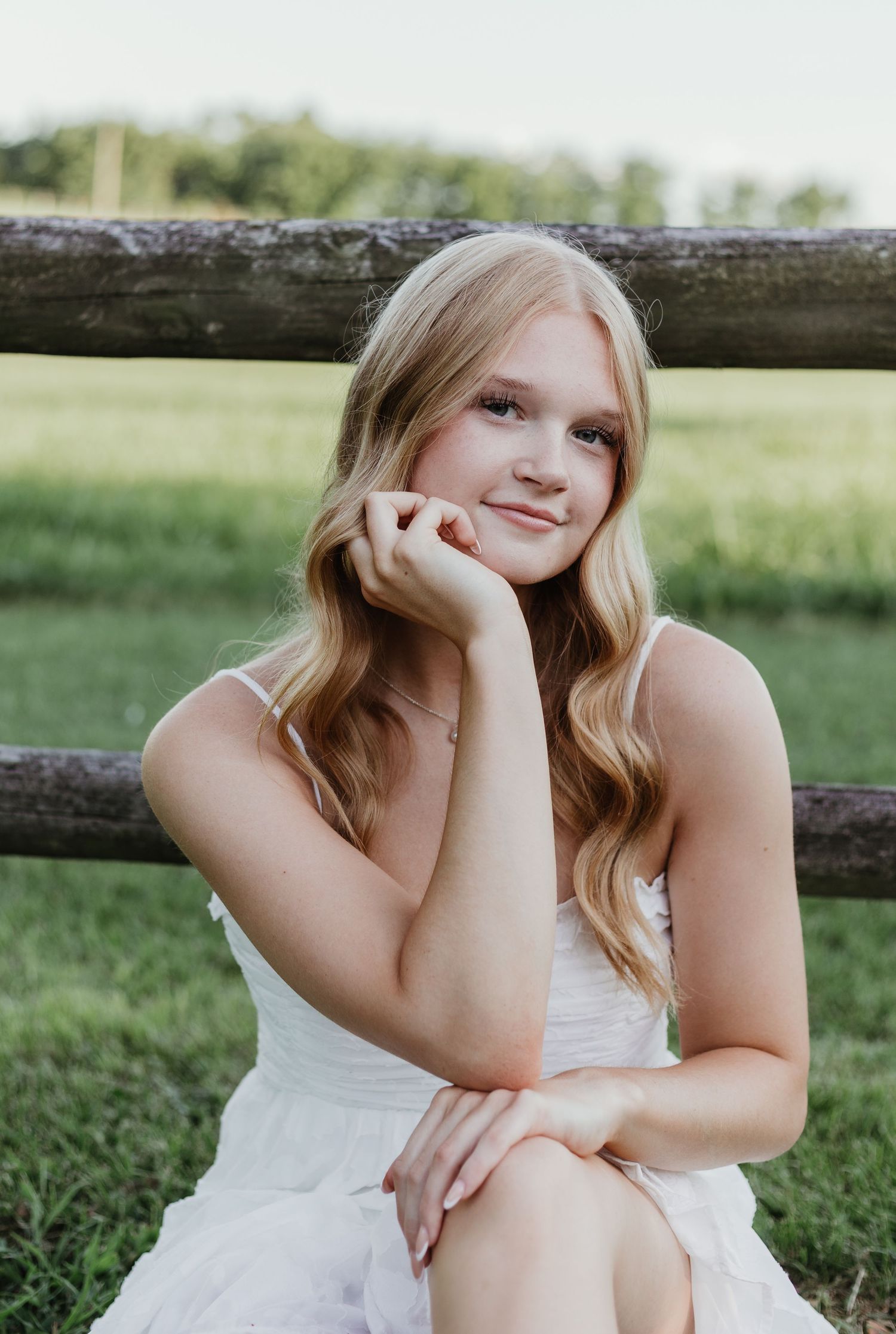 Senior in white dress sitting by rustic wooden fence in sunny outdoor setting with long wavy blonde hair.
