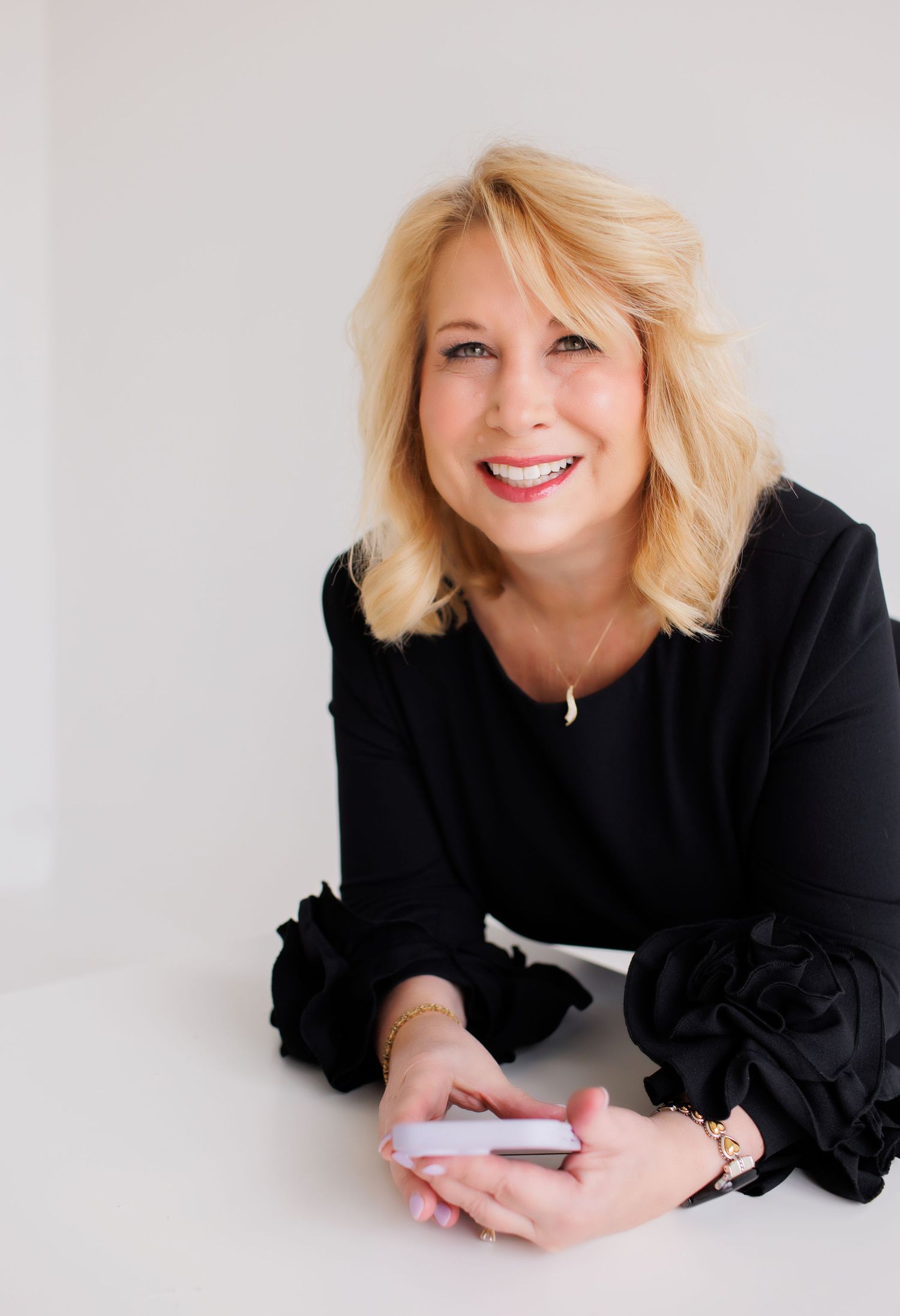 Smiling blonde woman in black ruffled top leaning on white surface holding a phone.