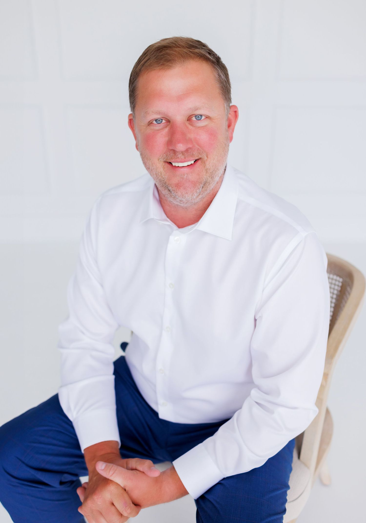 Professional man in white shirt and blue pants sitting confidently in a chair against a white background.