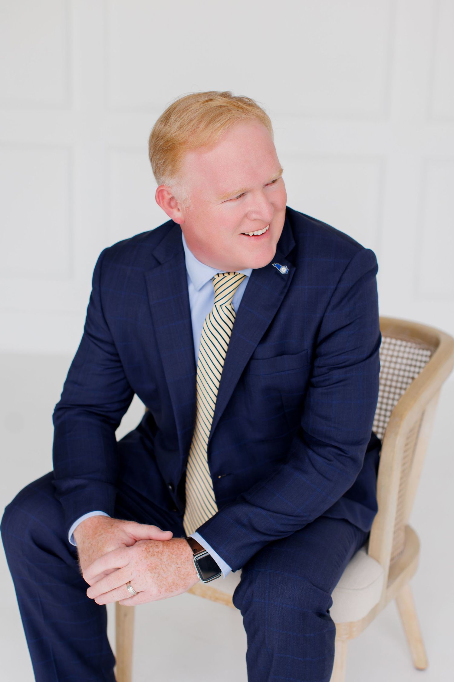 Man in navy suit and yellow striped tie sits smiling in a wooden chair against a white background.