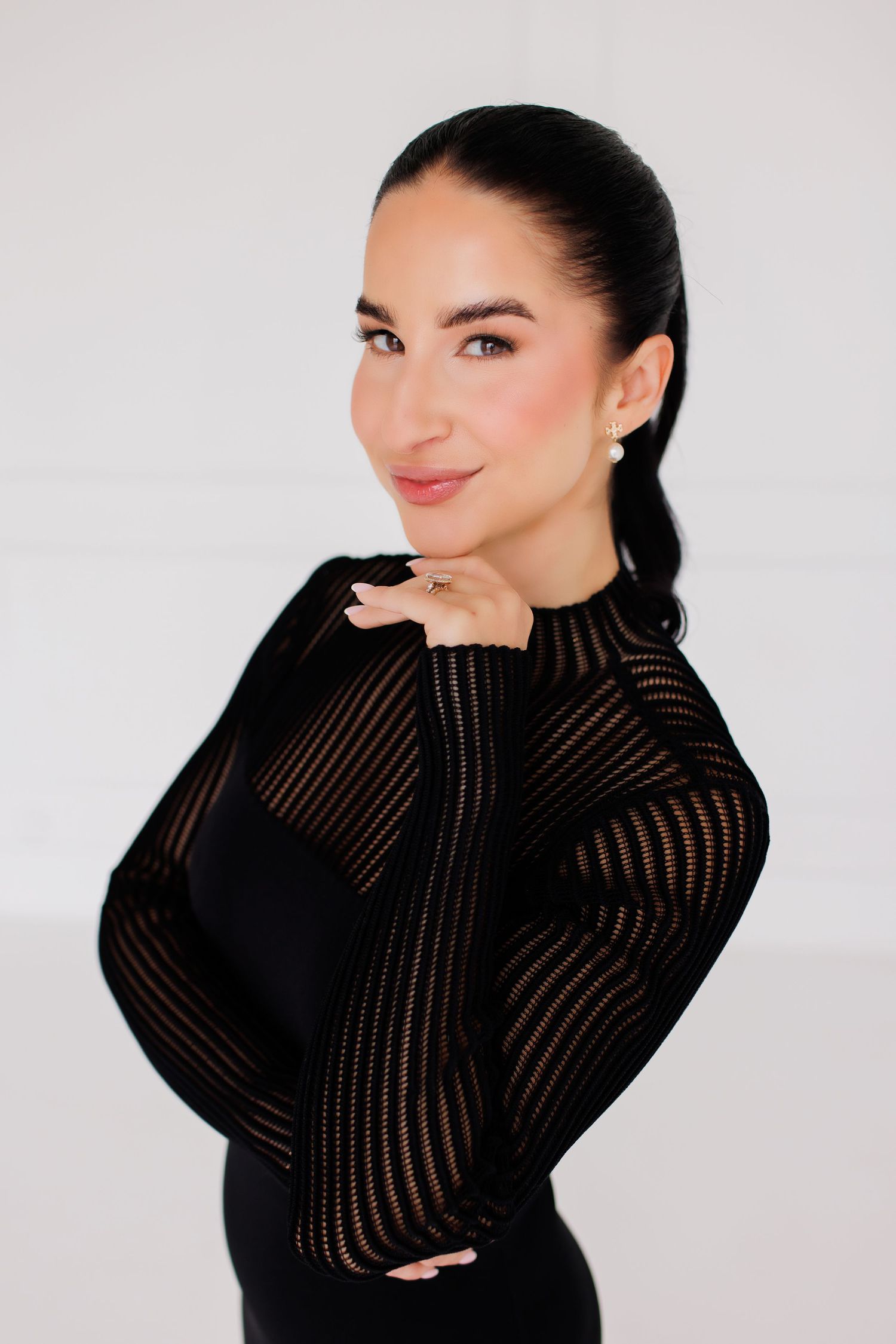 Woman in black sheer long-sleeve top posing confidently against white background with hand under chin.