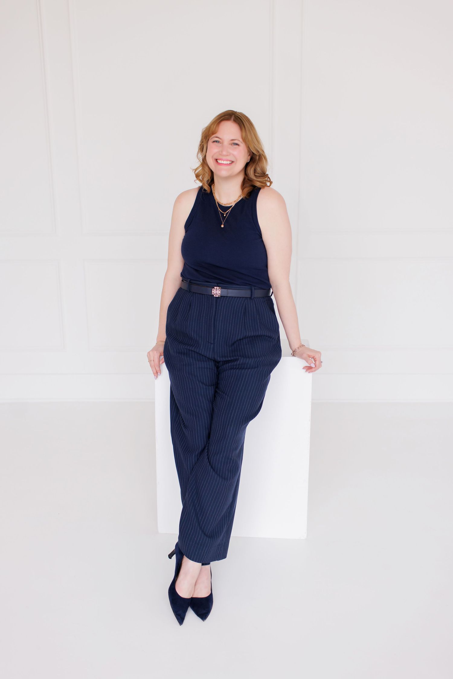 Woman in navy pinstripe trousers and sleeveless top leaning against white cube in bright studio.