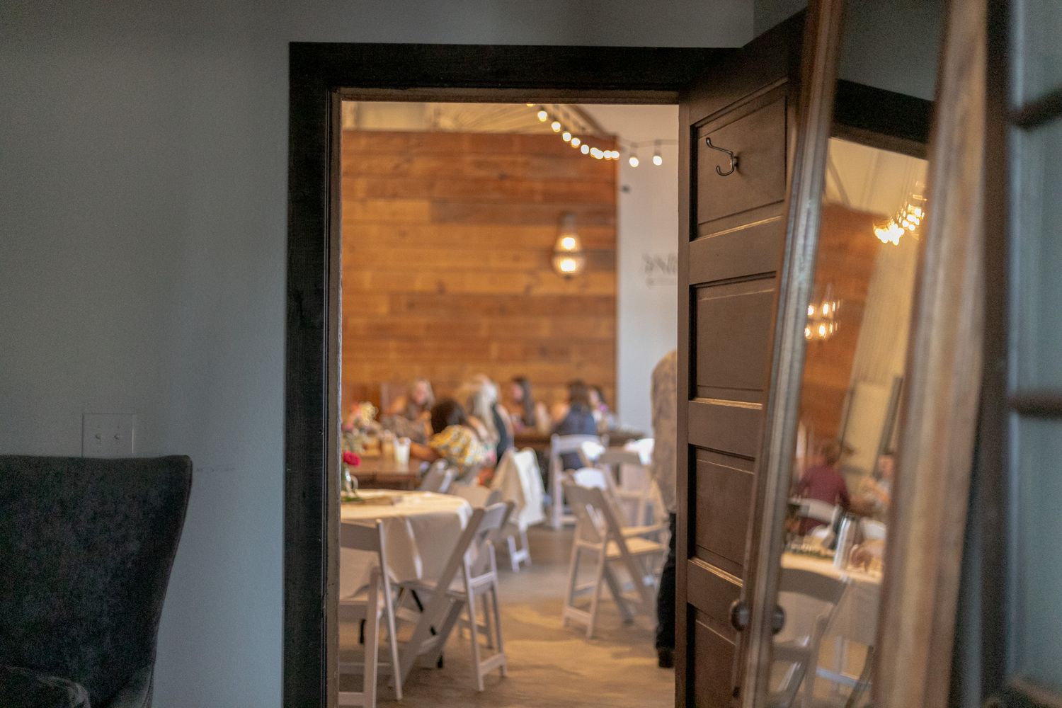 Warm interior view through an open wooden door into a decorated event dining room with string lights.