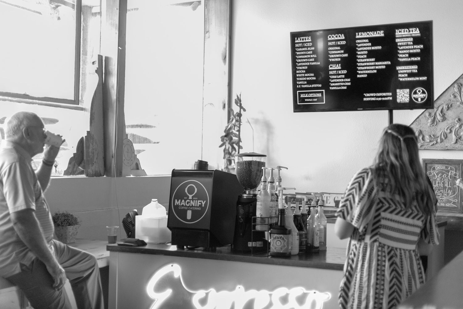 Black and white photo of customers at a coffee shop counter with an espresso menu board overhead.