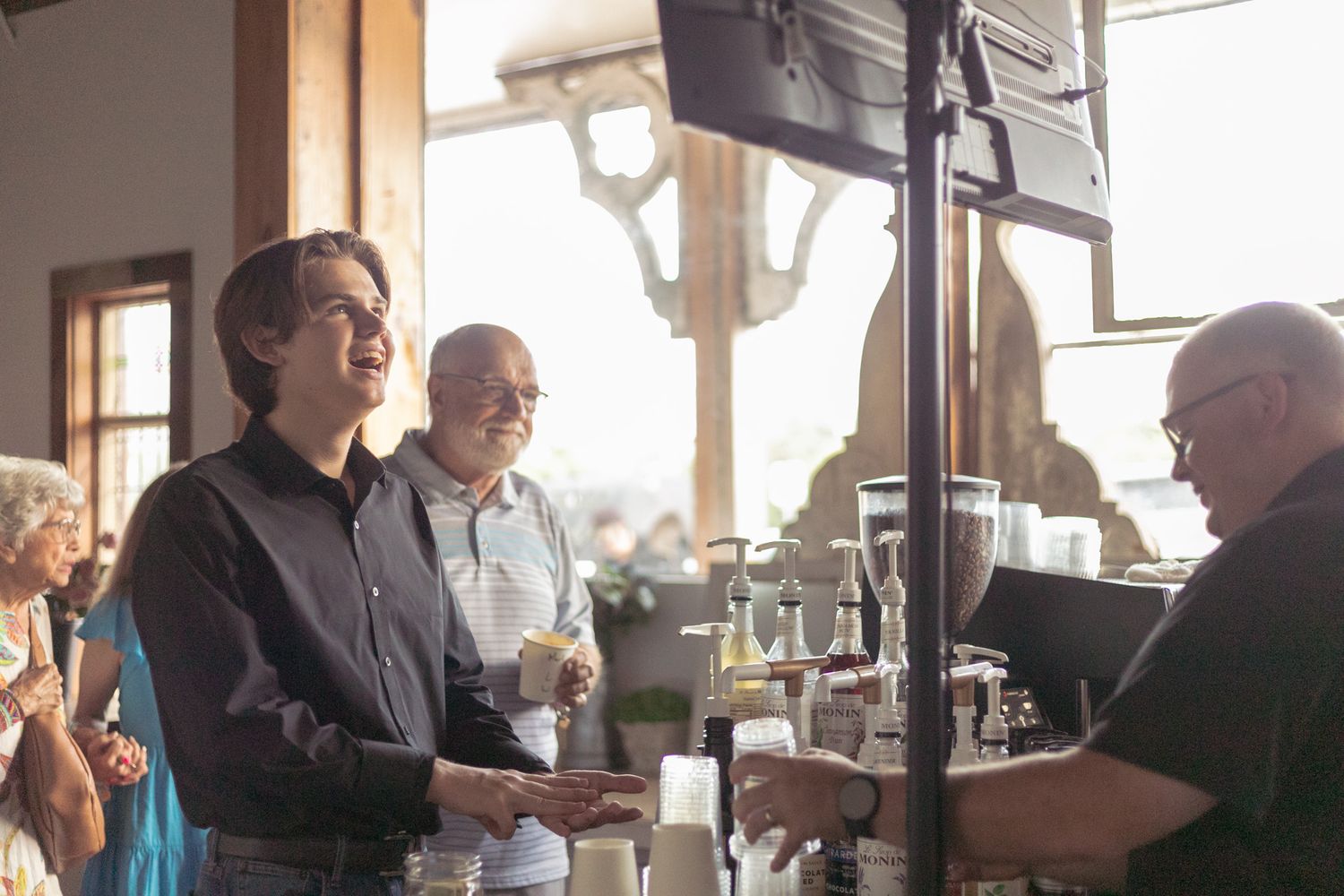 Smiling young man receiving a drink at a sunlit bar counter surrounded by guests at an event.