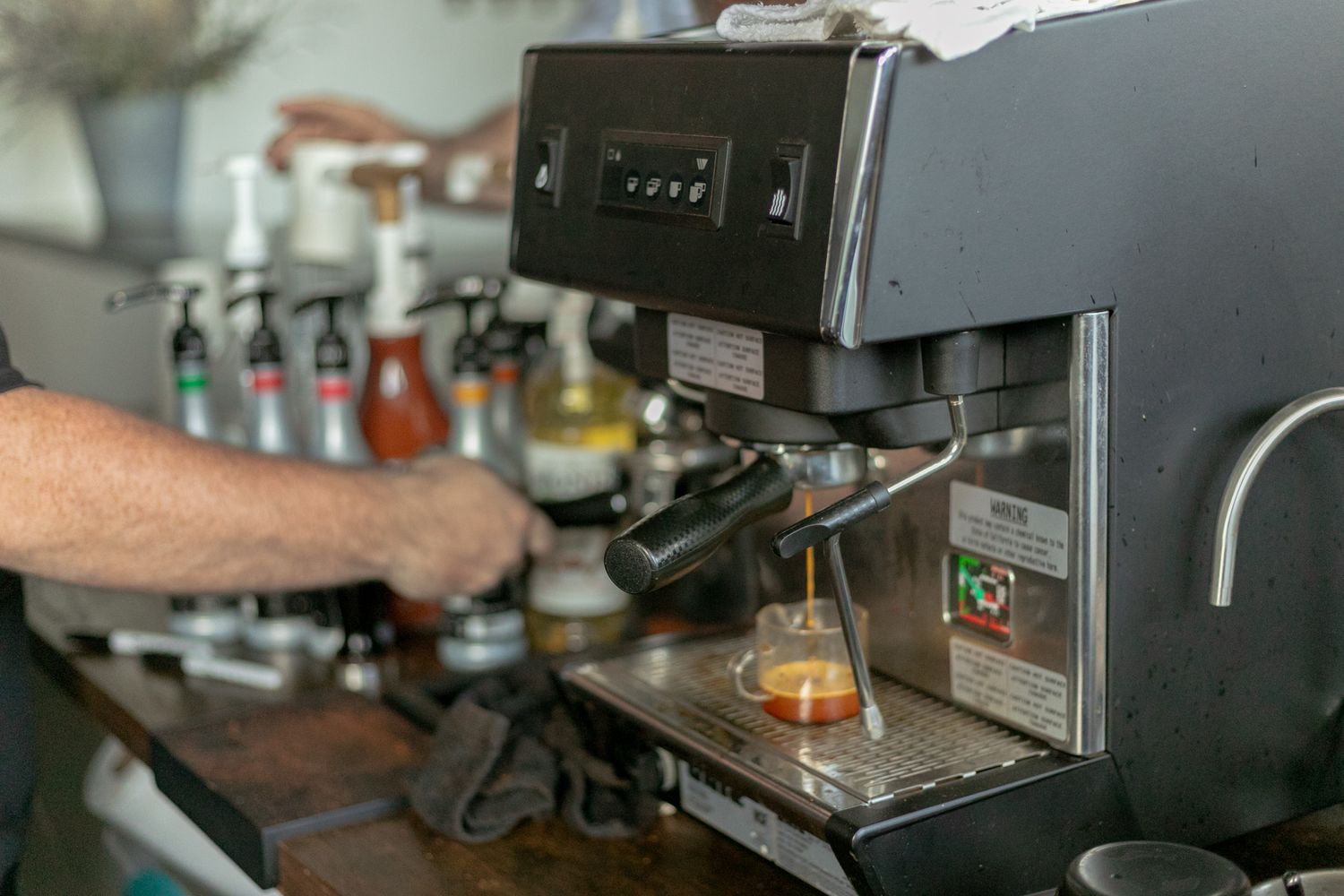 Barista operating a professional espresso machine with colorful syrups on a wooden bar counter.