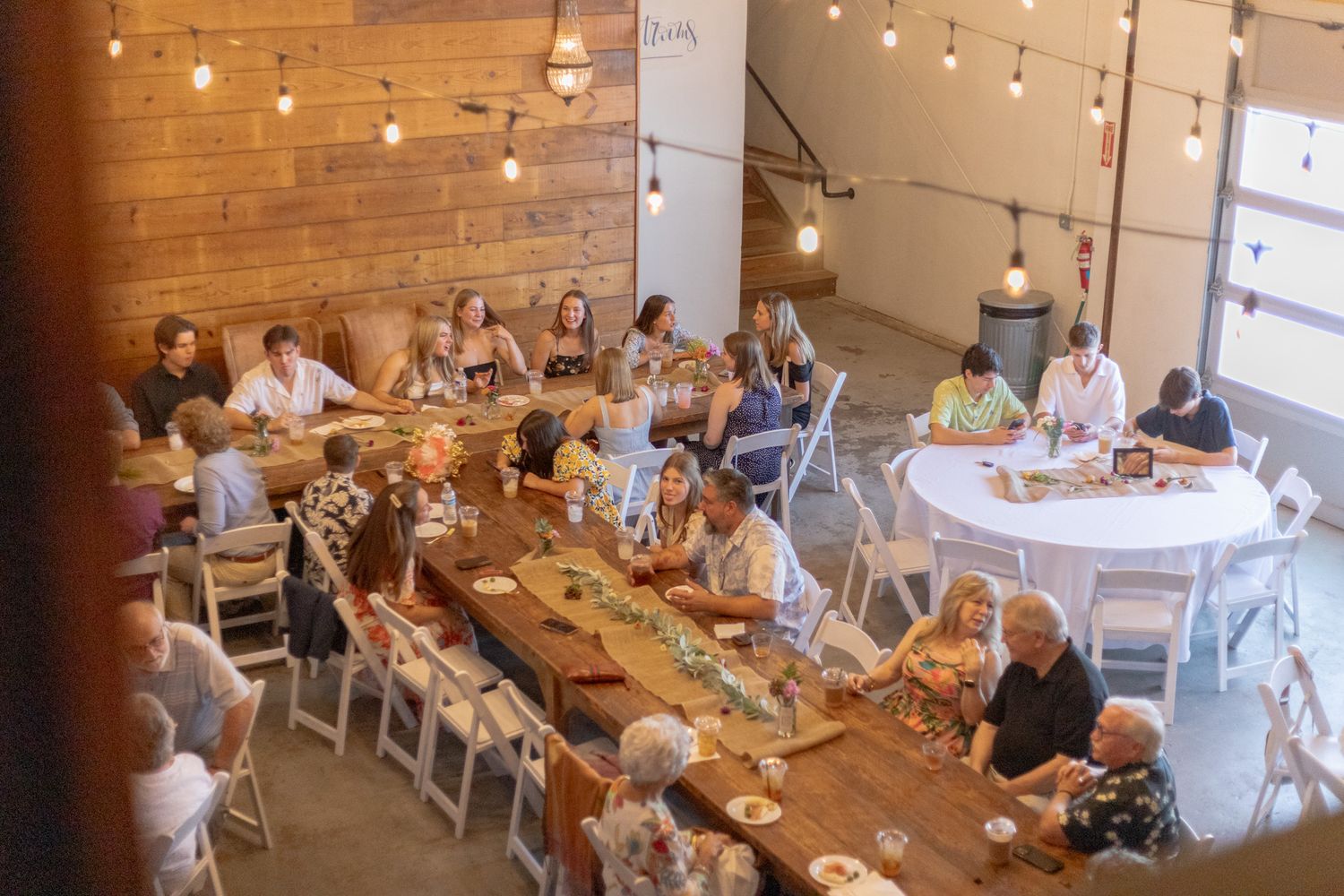 Guests dining at long wooden tables in a rustic venue with string lights and wood-paneled walls.