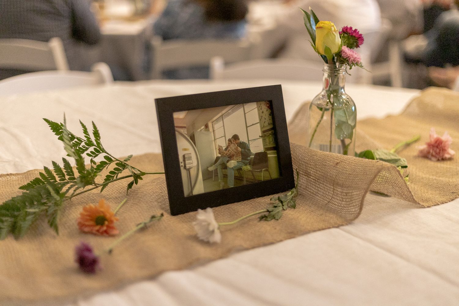 Framed photo centerpiece on burlap table runner surrounded by wildflowers and greenery.