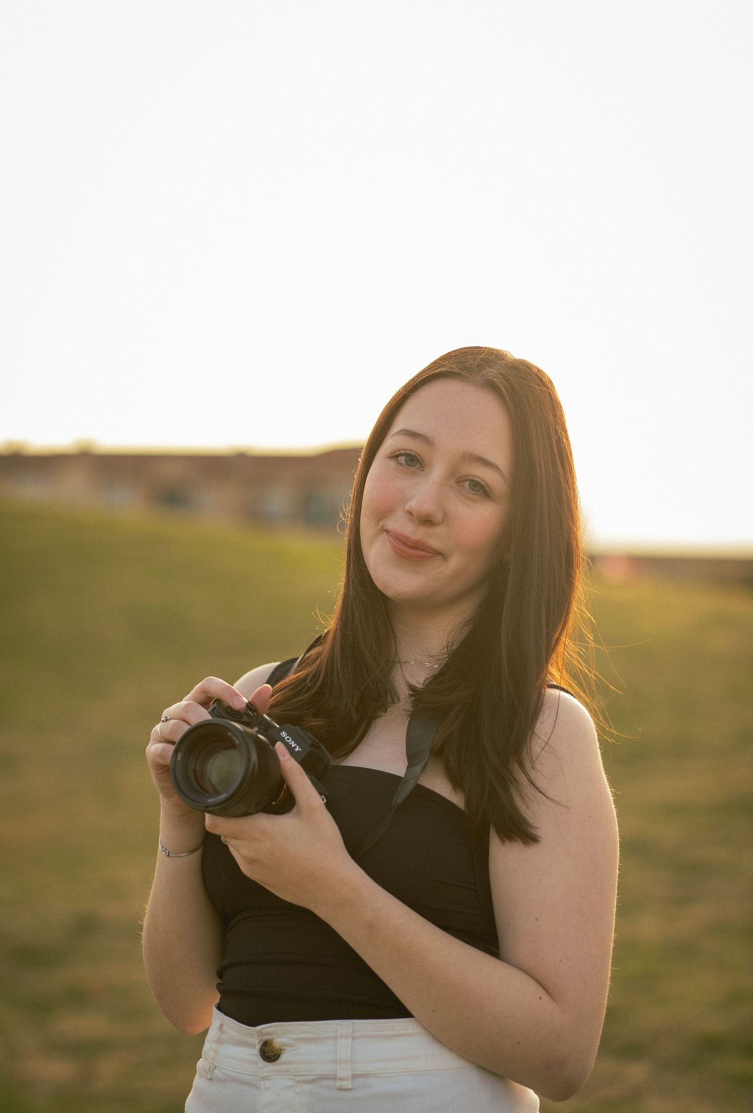 Young female photographer holding DSLR camera outdoors at sunset in black top and white pants.