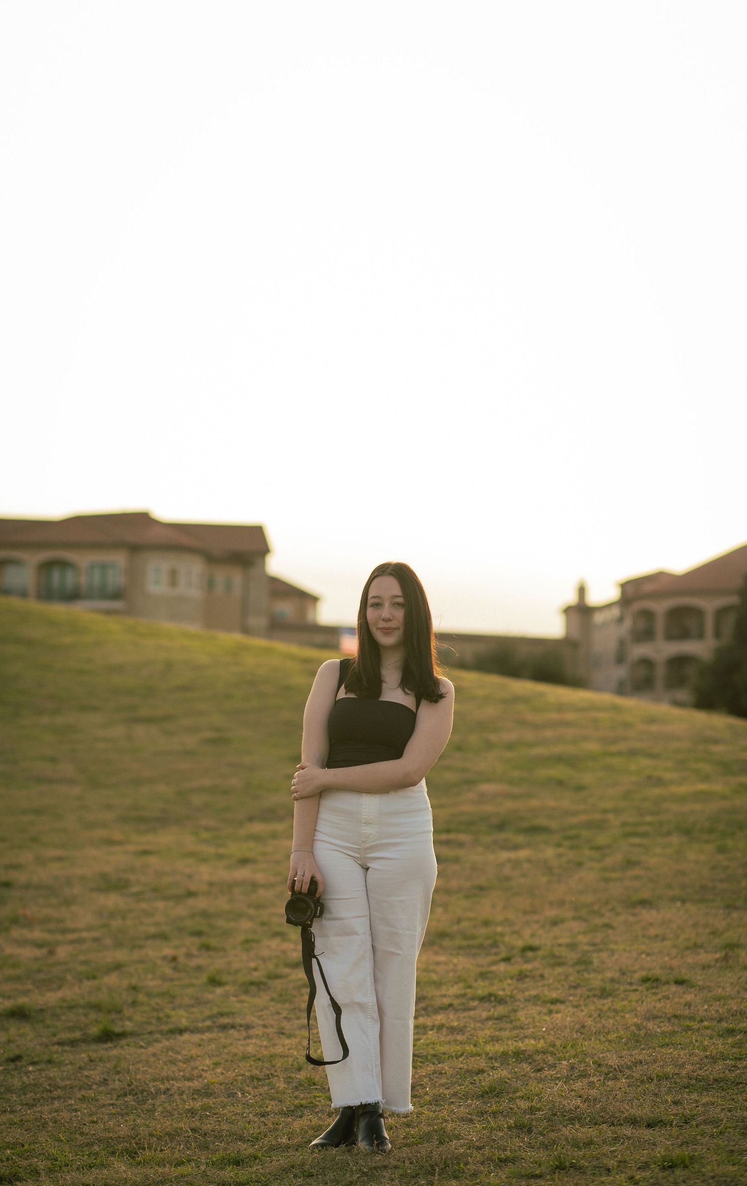 Young woman photographer standing in golden hour sunlight holding camera in white pants and black top.