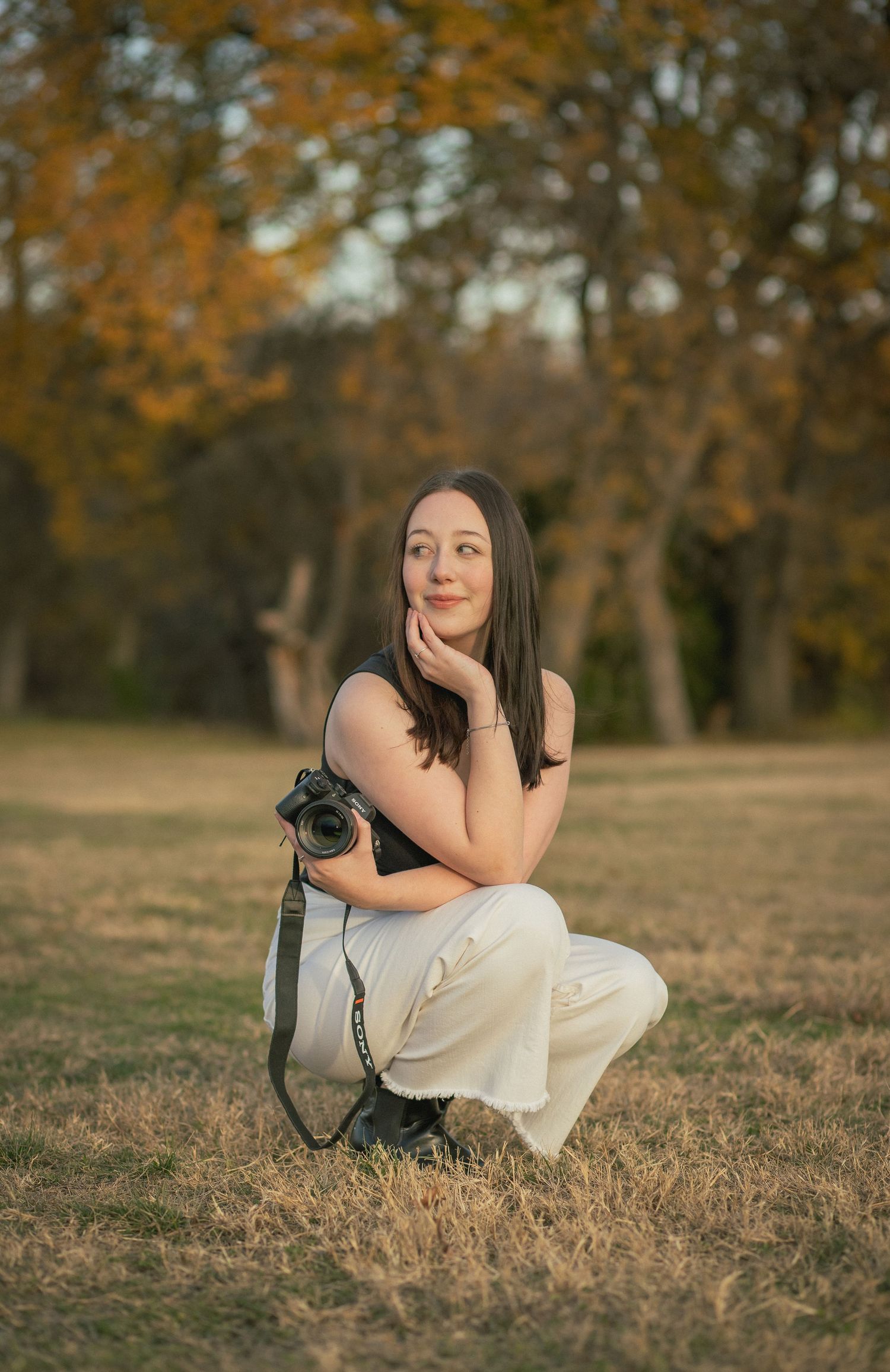 Female photographer crouching in autumn park holding DSLR camera, wearing black top and white pants.