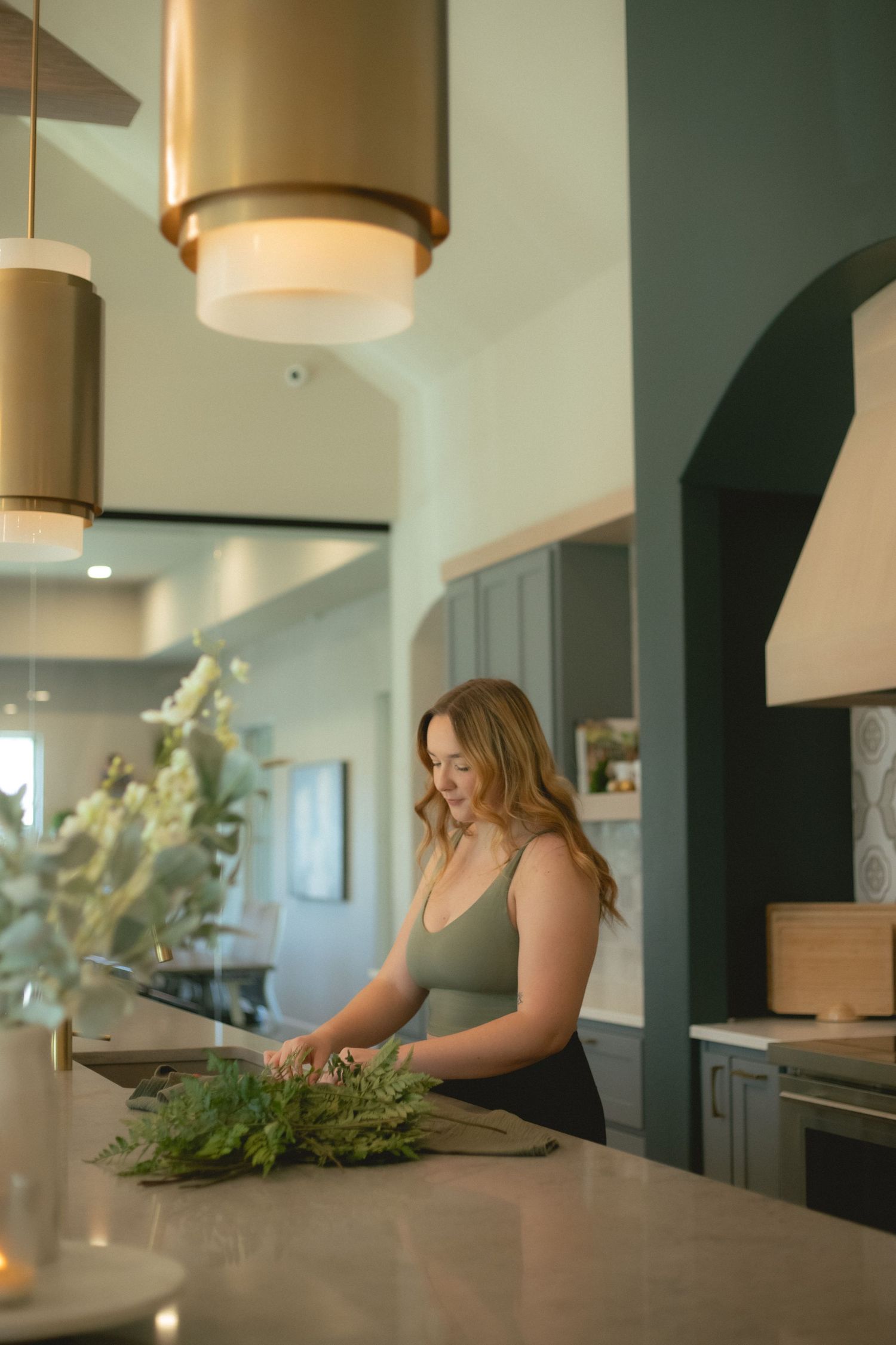 Woman arranging fresh greenery on kitchen counter under gold pendant lights in modern home.