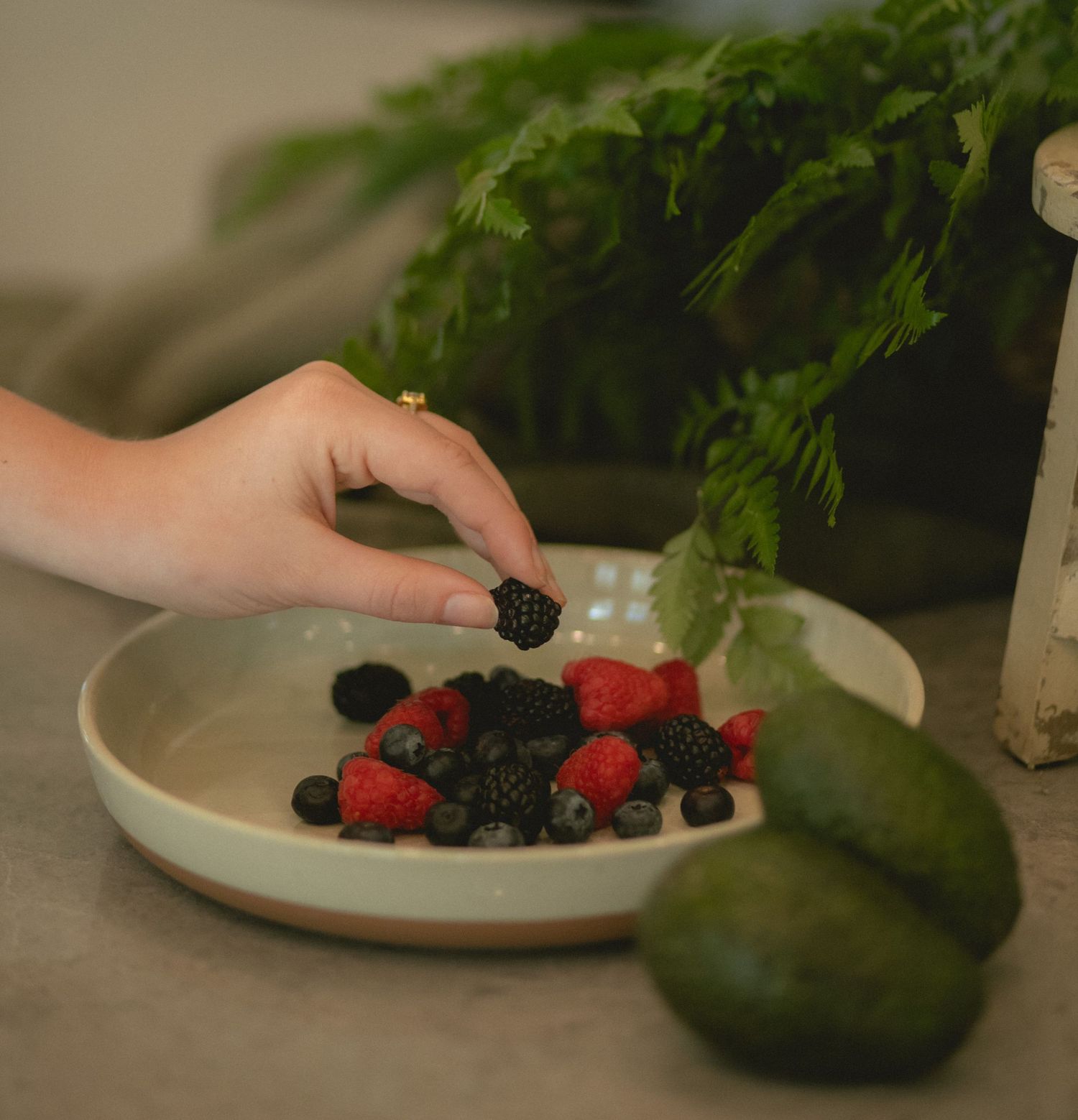 Hand picking a blackberry from a ceramic bowl of mixed berries next to avocados on counter.