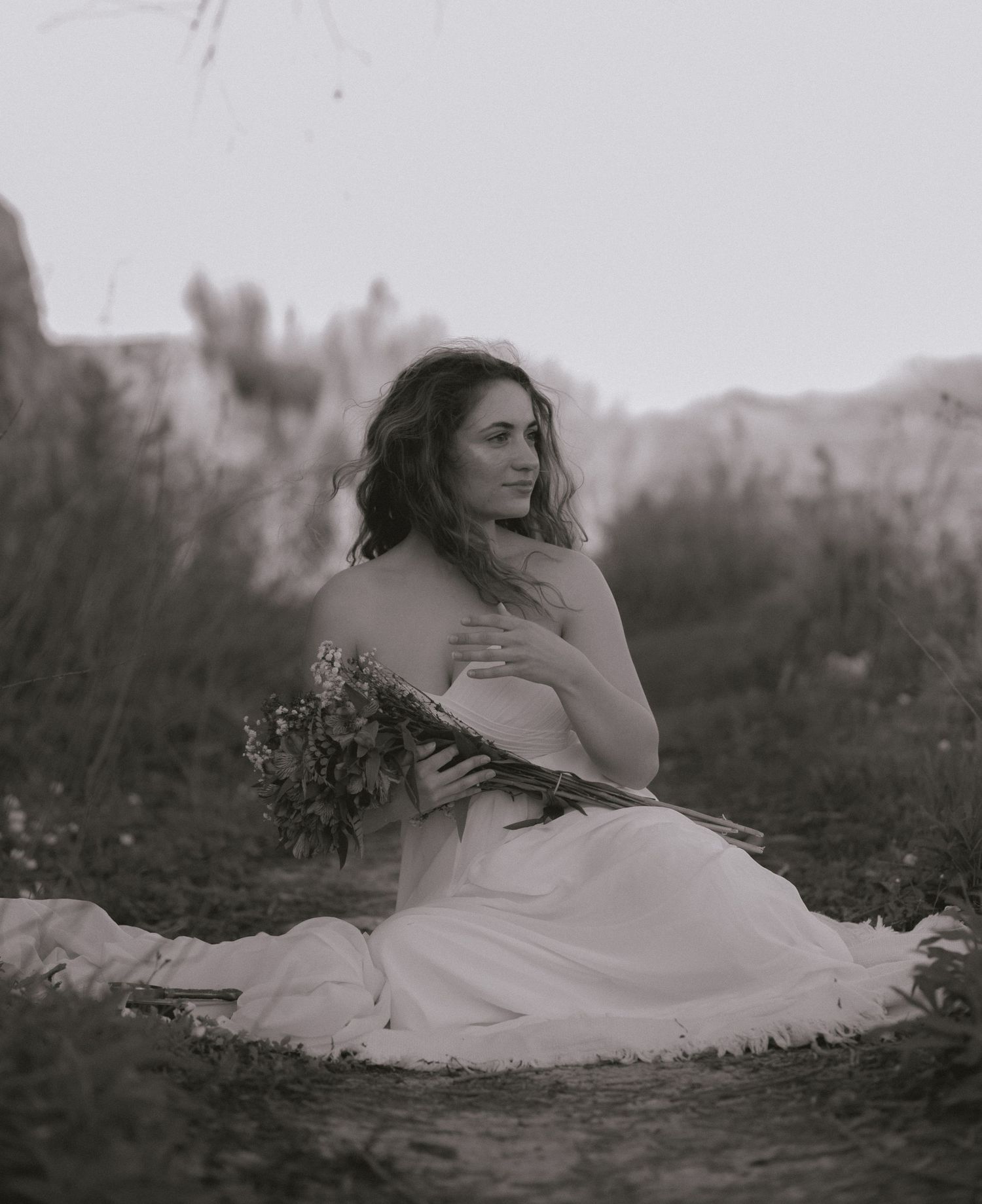 Black and white photo of a bride sitting outdoors holding a wildflower bouquet in a flowing white gown.