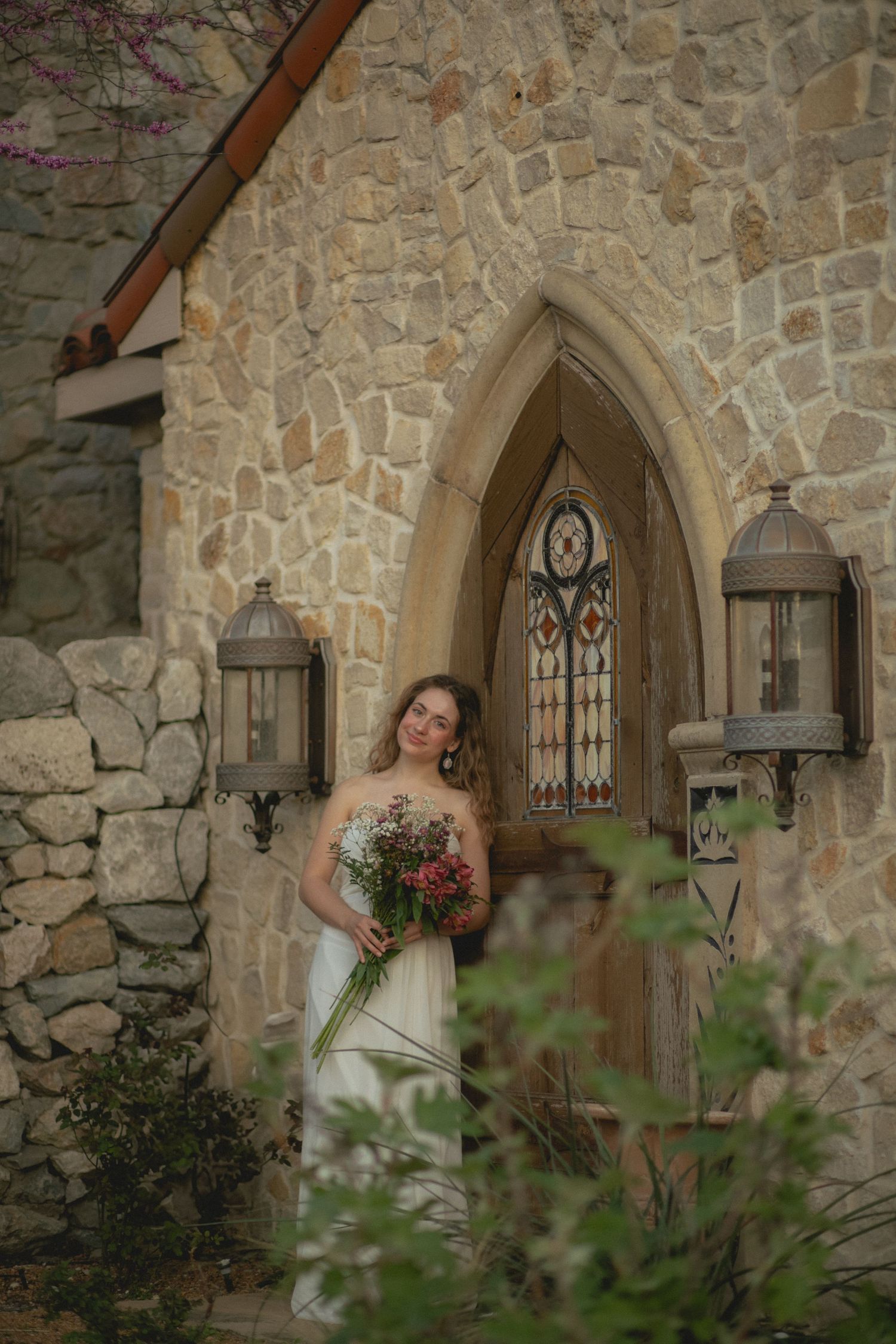 Bride in white strapless gown holding bouquet stands before a stone chapel with arched stained glass door.