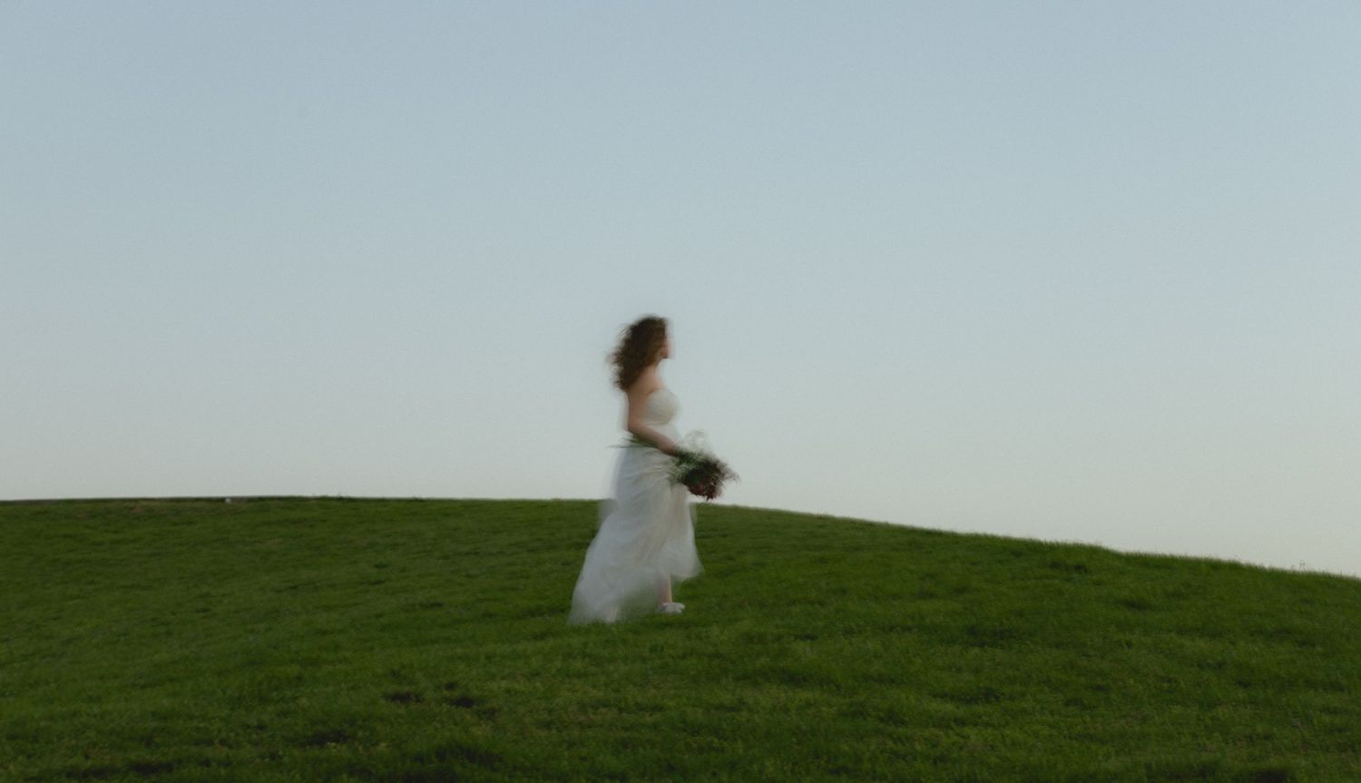 Bride in white flowing gown walks across a lush green hill against a clear blue sky.