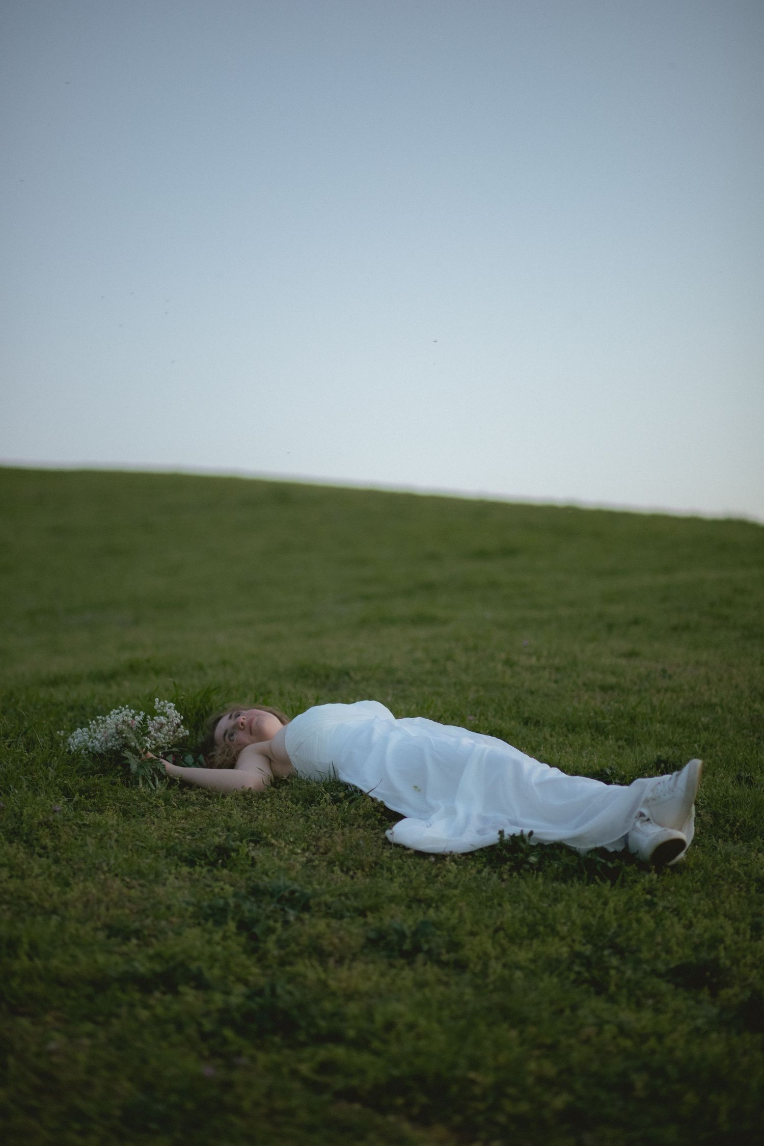 Bride in white gown lies peacefully on green grass holding a small floral bouquet.