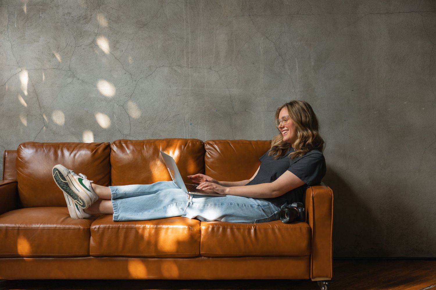 Woman relaxing on brown leather sofa with laptop, smiling in stylish modern living room with concrete wall.