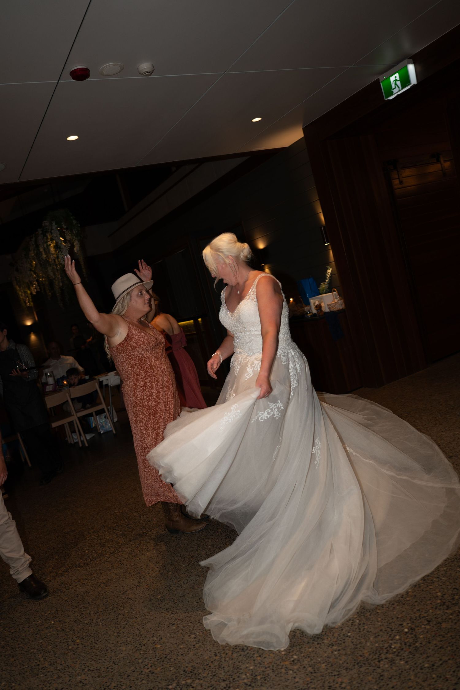Bride in white lace gown dancing joyfully with a guest in a hat at a wedding reception.