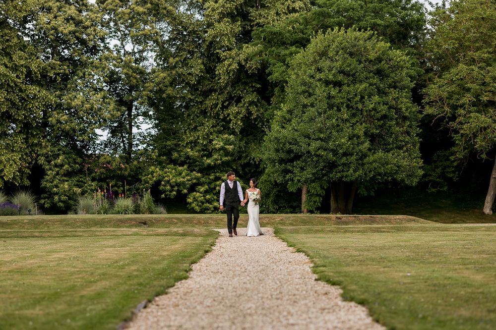Bride and groom walk hand in hand along a gravel path surrounded by lush green trees and manicured lawns.