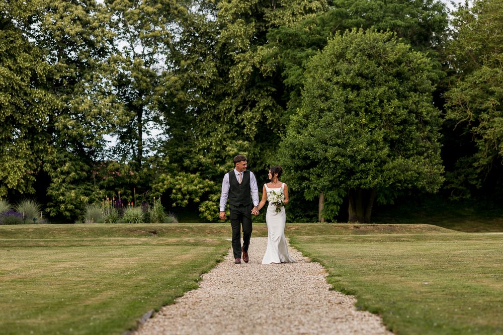 Bride and groom walking hand in hand along a gravel path surrounded by lush green trees.