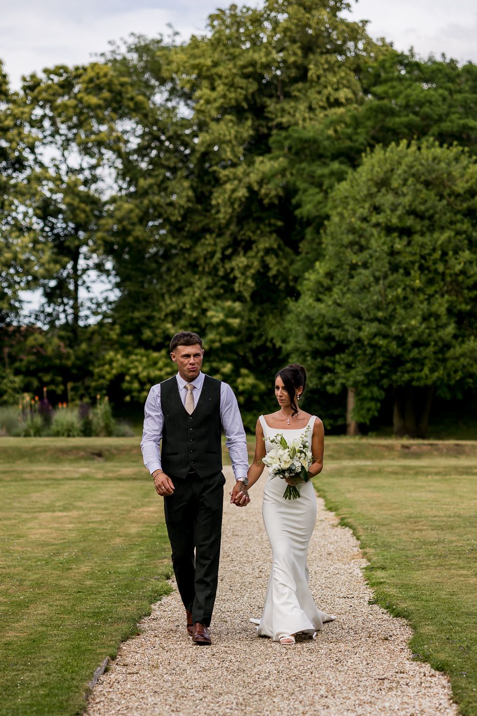 Bride and groom walk hand-in-hand along a gravel path surrounded by lush green lawns and trees.