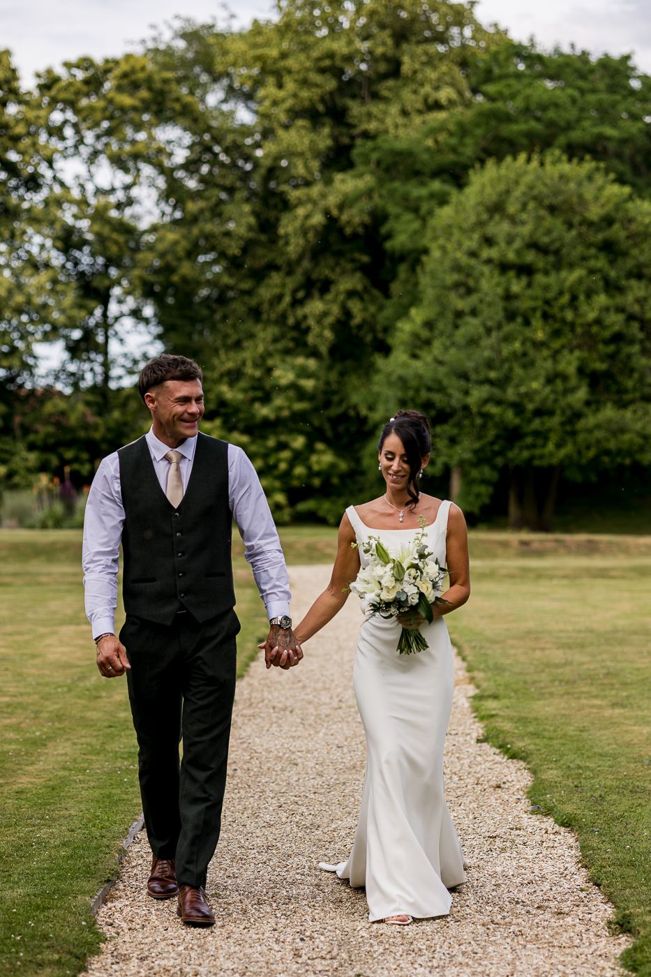 Bride and groom walk hand-in-hand on a gravel path, she holds a white bouquet, surrounded by lush greenery.