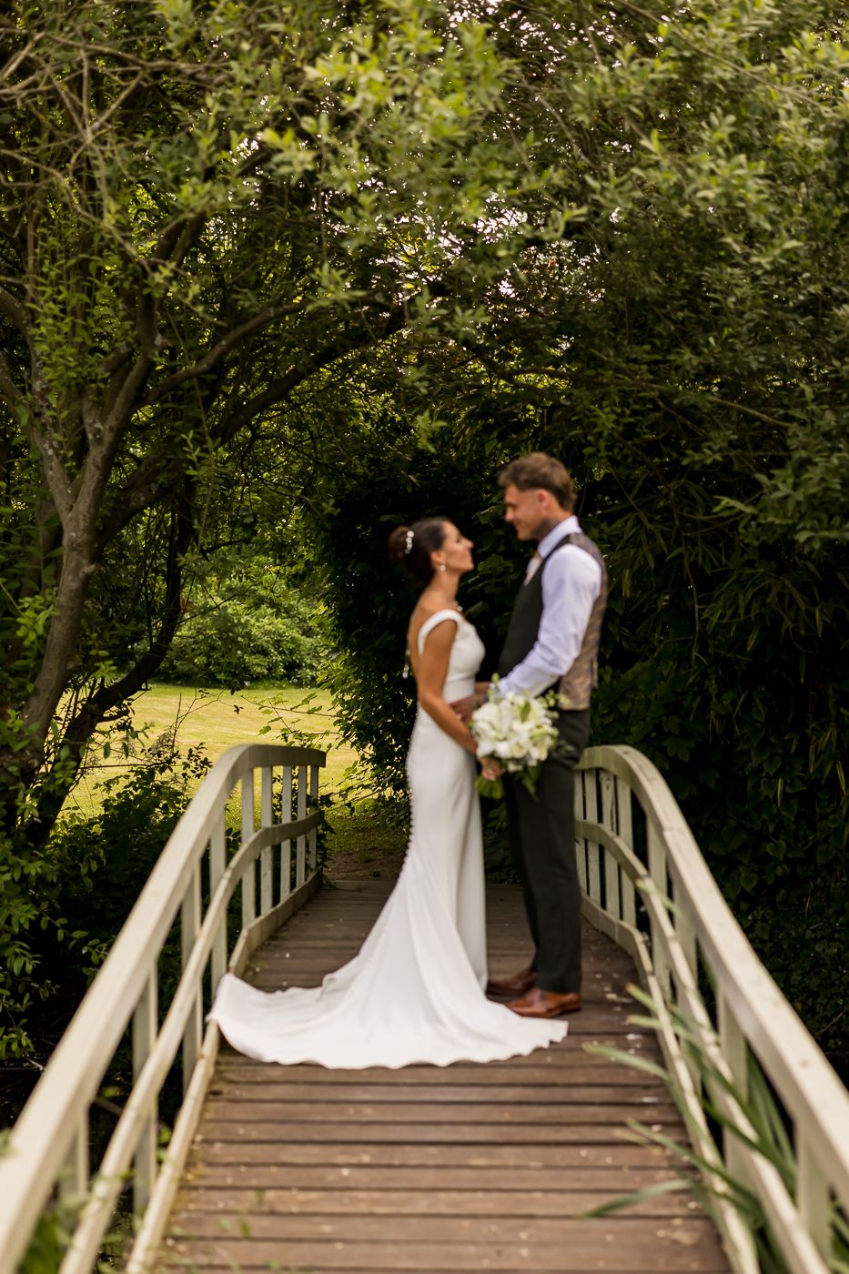 Bride and groom share a romantic moment on a wooden bridge surrounded by lush green trees on their wedding day.