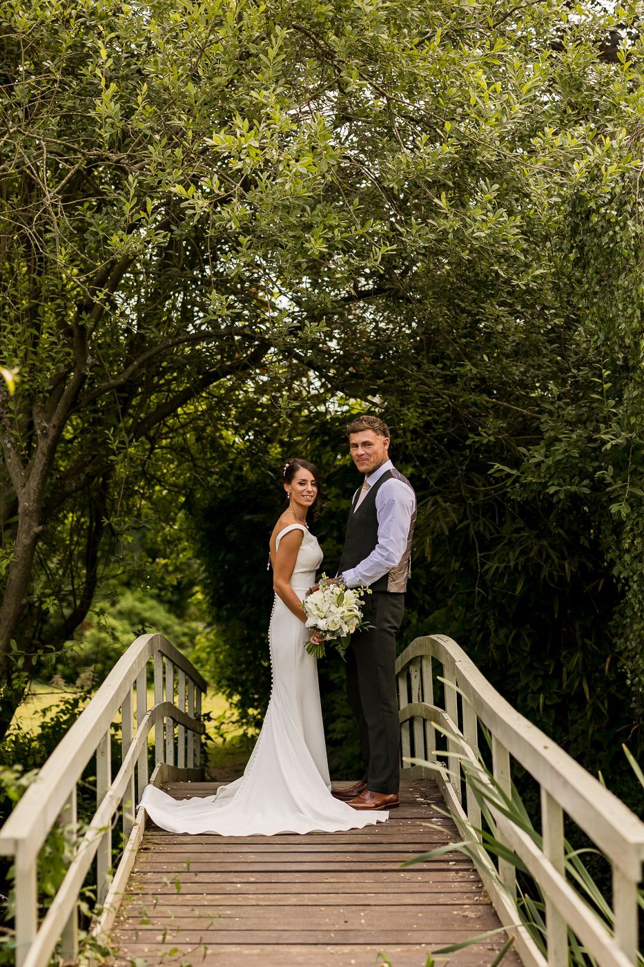 Bride and groom pose on a wooden bridge surrounded by lush green trees in a romantic garden wedding portrait.