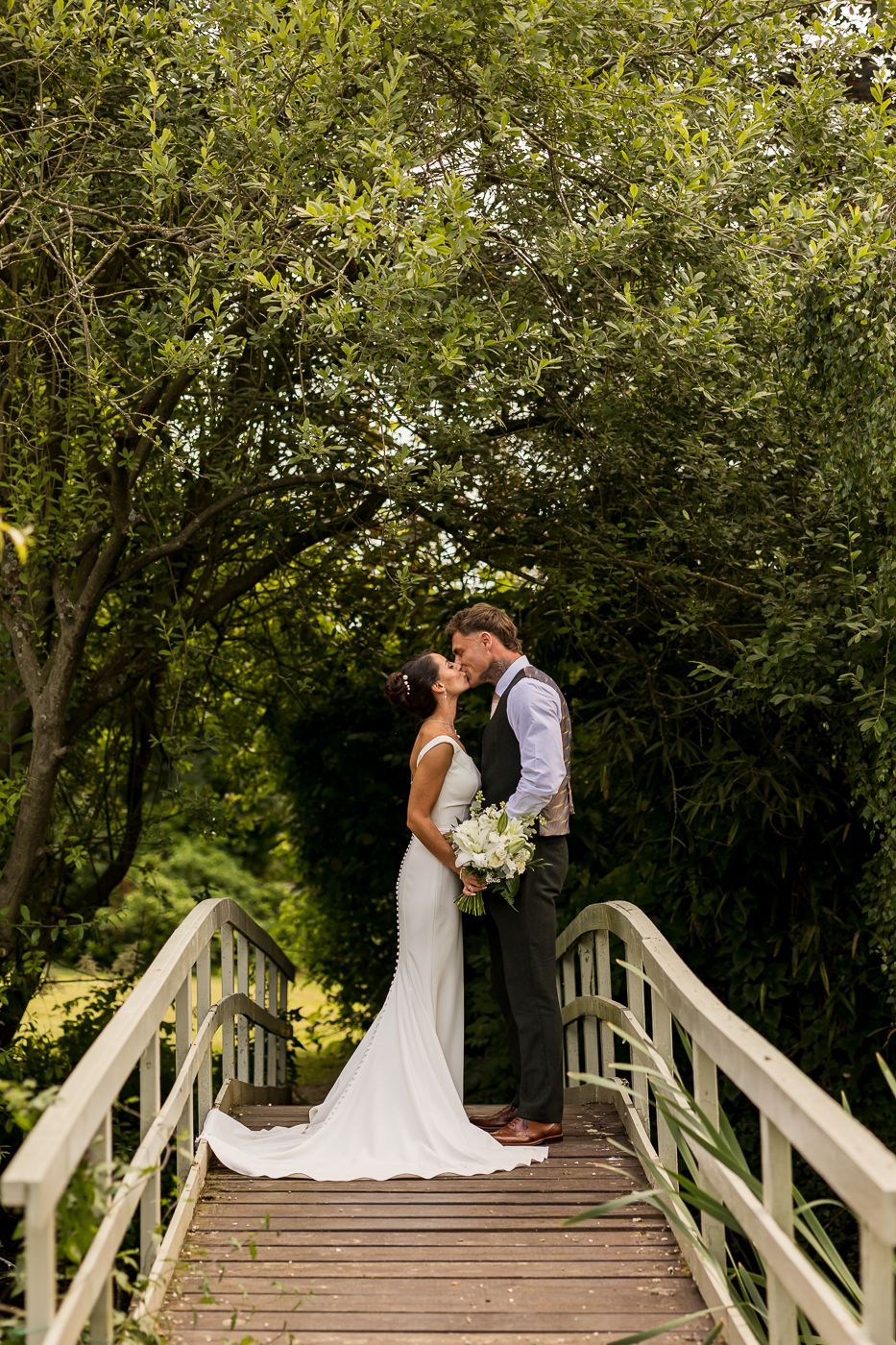 A bride and groom share a romantic kiss on a wooden bridge surrounded by lush green trees on their wedding day.