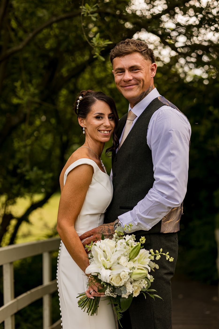 A smiling bride in a white dress holds a bouquet of white flowers beside a groom in a grey vest outdoors.