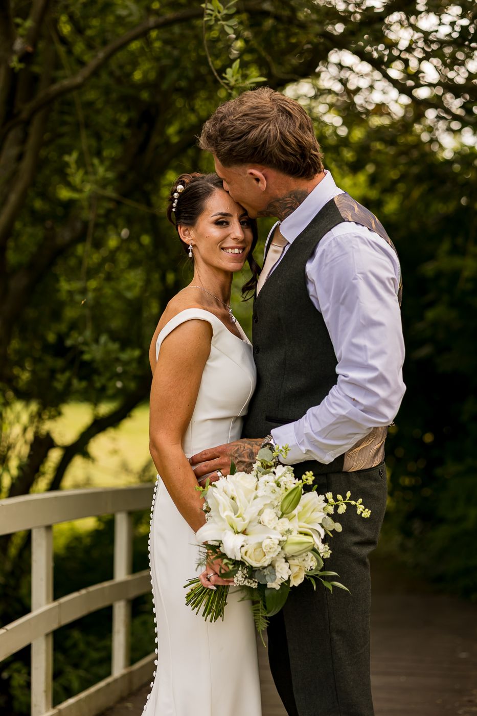 A bride holding white flowers smiles as her groom kisses her cheek on a wooden bridge surrounded by lush greenery.