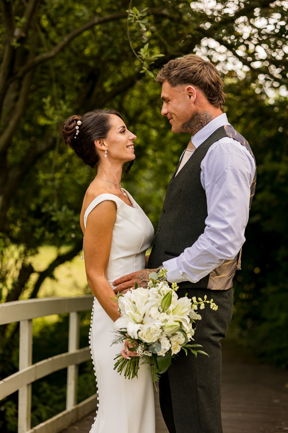 Bride and groom share a loving gaze on a garden bridge, bride in white gown holding a white floral bouquet.