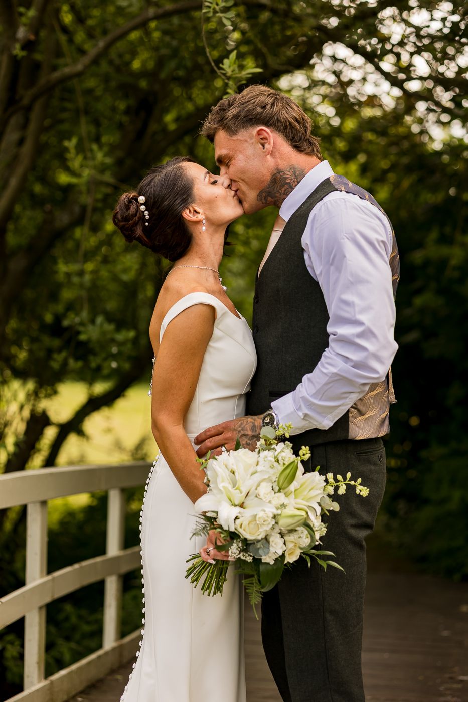 Bride and groom share a romantic kiss on a garden bridge, bride holding white floral bouquet in elegant wedding attire.